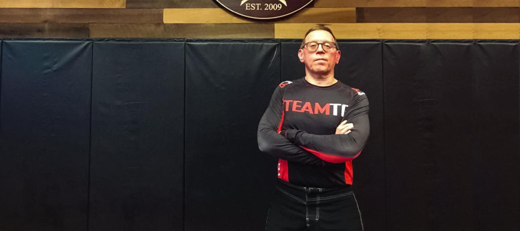 Laszlo Ember posing with arms crossed under the logo at the BJJ school